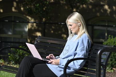 A student on campus sits outside on a bench looking at their laptop.